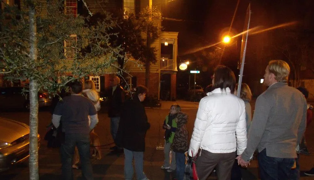 A group of people gathers on a street corner at night some engaging in conversation others walking by near a tree with a streetlight in the background