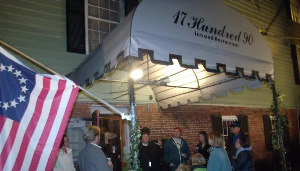 A group of people gather outside the 17 Hundred 90 Inn and Restaurant beneath an awning with an American flag prominently displayed in the foreground