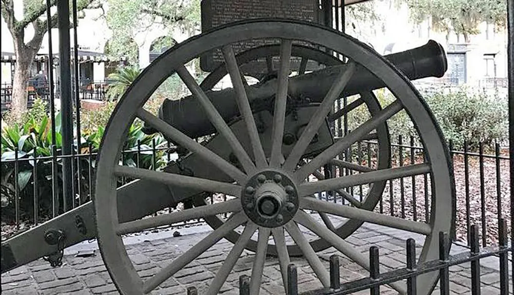 An old cannon mounted on large wooden wheels is displayed behind a metal fence possibly in a historical or public park setting