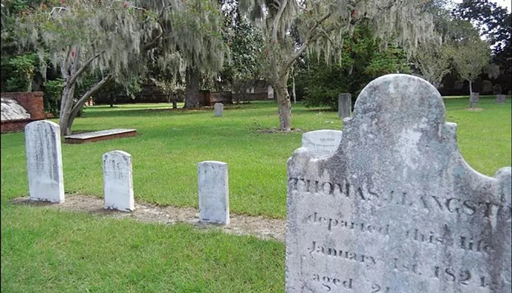 The image shows a tranquil cemetery scene with weathered gravestones and Spanish moss hanging from trees in the background