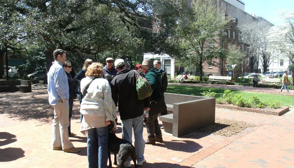 A group of people one with a dog appears to be engaged in a conversation or a tour in an outdoor setting with trees benches and buildings in the background