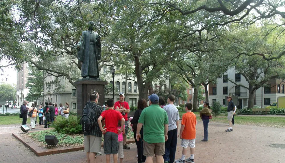 The image depicts a group of people gathered around a tour guide in a park with a statue and large trees in the background