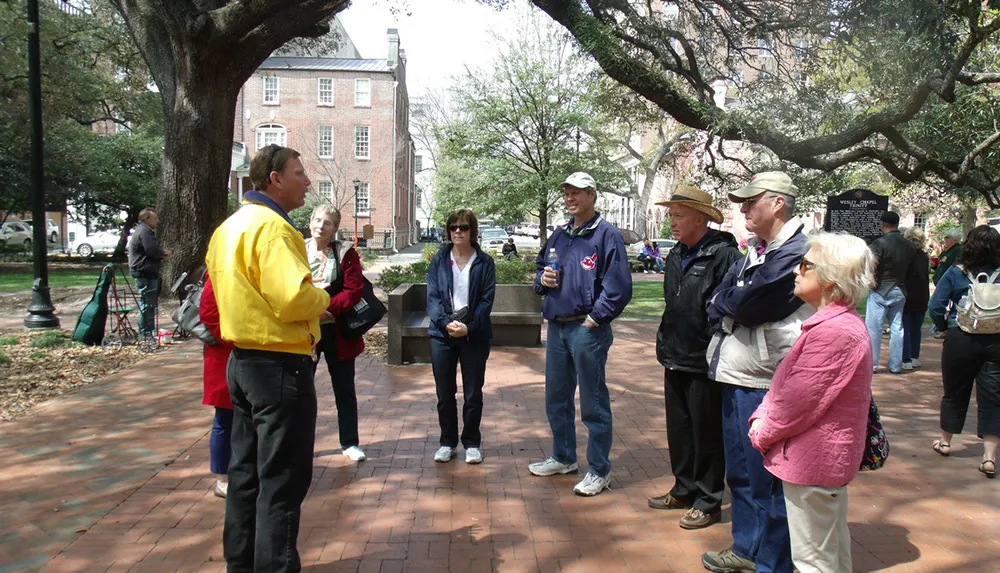 A group of people are attentively listening to a person in a yellow jacket possibly a tour guide in an outdoor setting with trees and historic buildings in the background