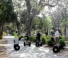 A group of six people are posing with Segways in front of vibrant pink flowers and Spanish moss-draped trees under a clear sky