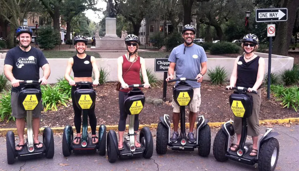 Five people wearing helmets are smiling for the camera while standing on Segways in a park-like setting