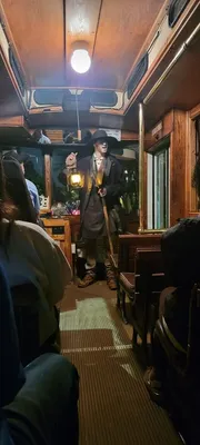 A person in a period costume holding a lantern stands inside a vintage train carriage, seemingly addressing the passengers.