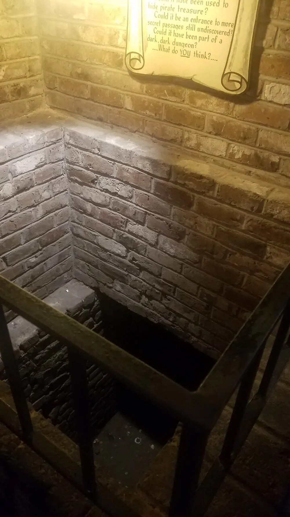 The image shows a dimly lit brick-lined staircase leading down to a dark opening with a plaque on the wall suggesting a historical or mysterious narrative about pirate treasure or secret passages