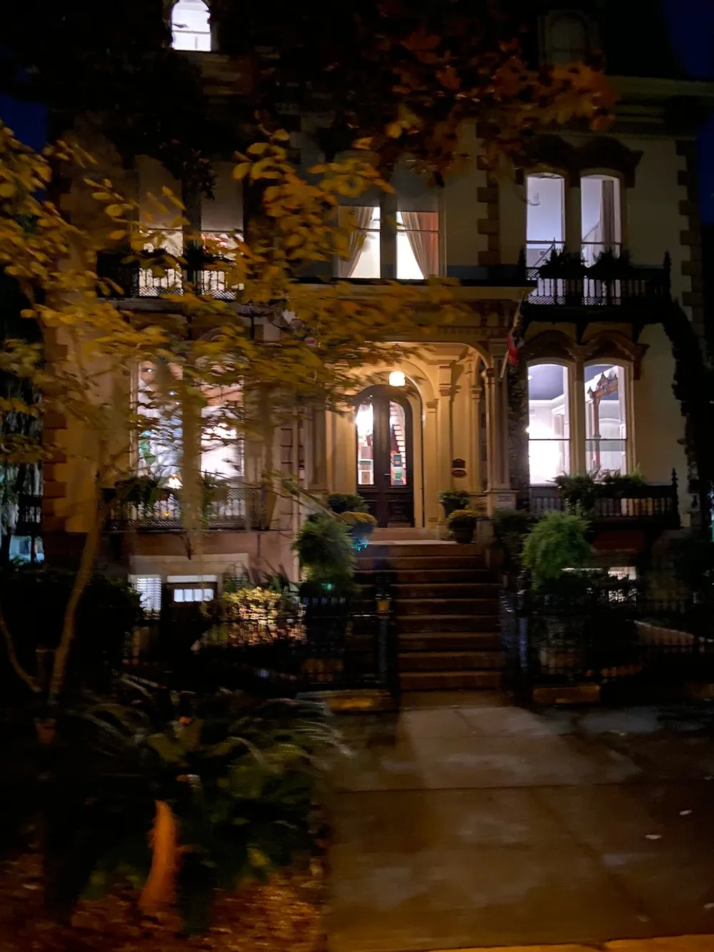 The image shows a warmly lit brownstone townhouse at night framed by foliage and featuring a classic stoop with iron railings