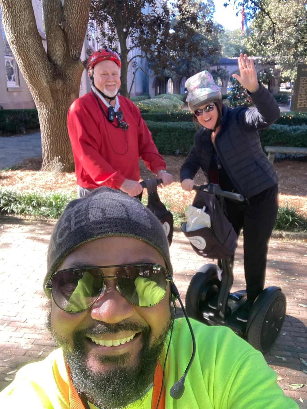 A cheerful group of three people one taking a selfie with two others behind him is enjoying a sunny day outdoors possibly on a Segway tour