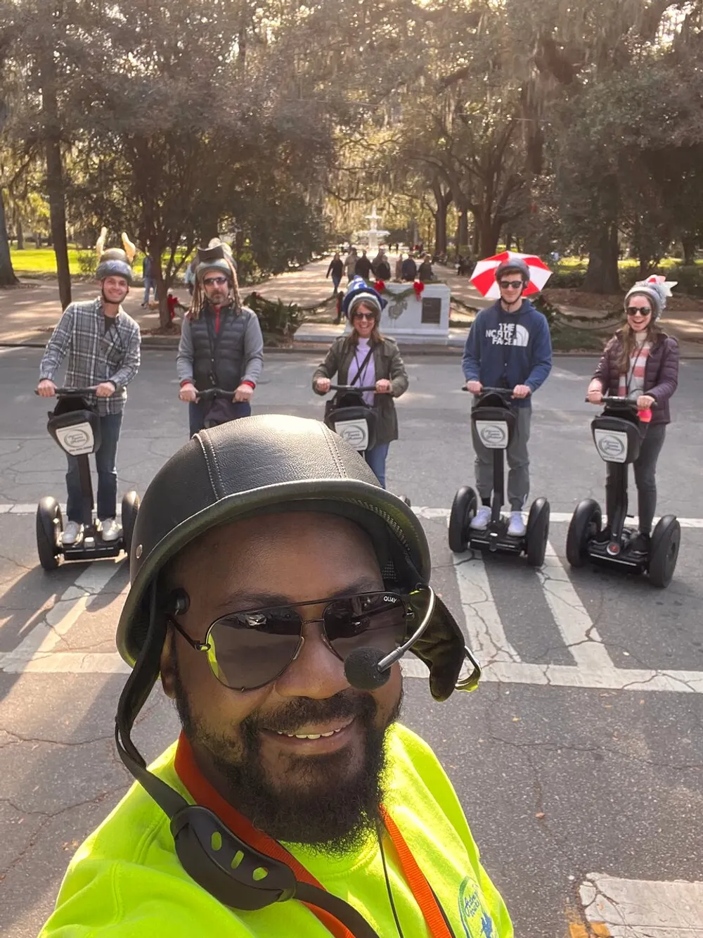 A smiling person in the foreground takes a selfie with a group of people behind them riding Segways in a park-like setting