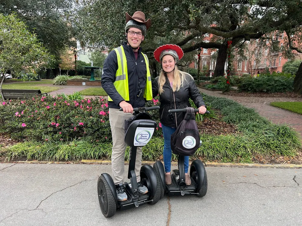A man and a woman both wearing helmets are standing with Segways in a park-like setting with greenery and a brick building in the background