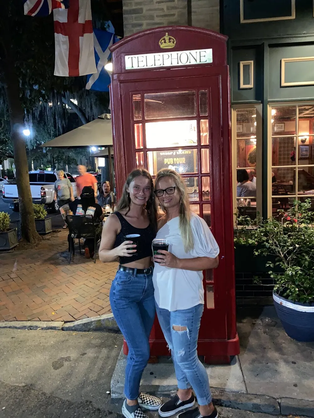 Two people are smiling for a photo in front of a red telephone box holding drinks with a backdrop featuring flags and a restaurant setting