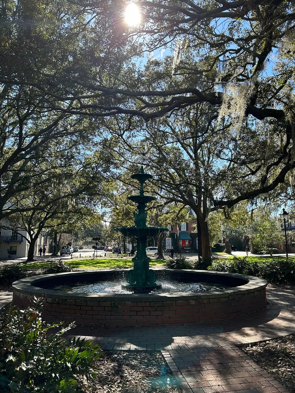 The image showcases a sunlit fountain surrounded by brick pavement under the shade of large trees draped with Spanish moss in a peaceful park setting