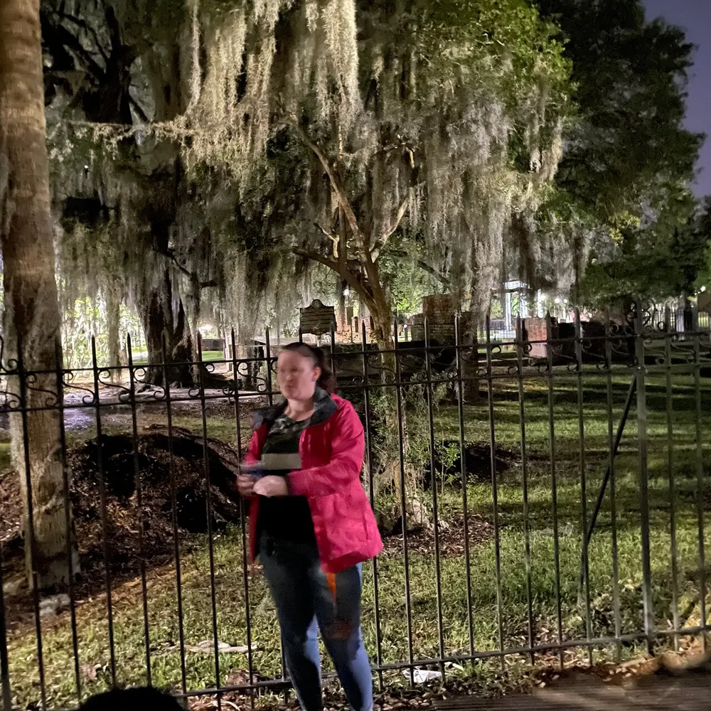 A person stands in front of a wrought iron fence at night with Spanish moss-draped trees and tombstones in the background suggesting a graveyard setting