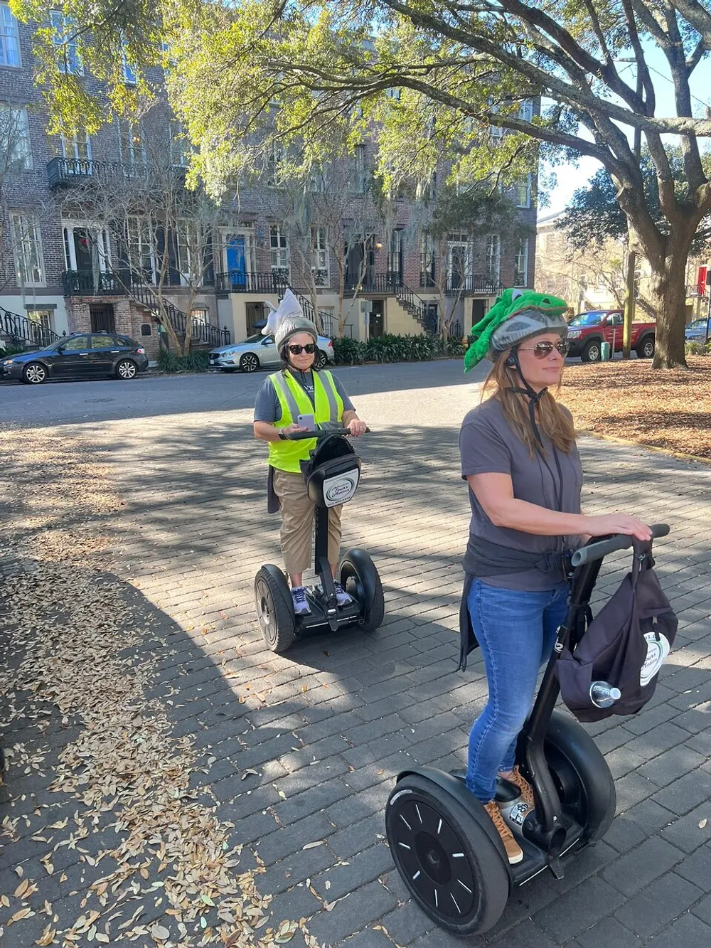 Two people one wearing a helmet with a quirky alligator design and the other sporting a vest are riding Segways on a tree-lined path with historic buildings in the background