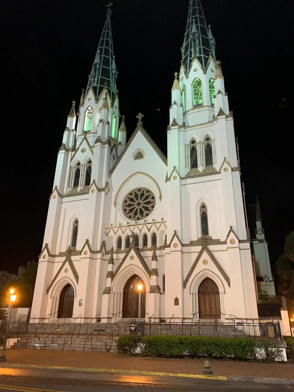 The photo depicts a grand white gothic revival-style church captured at night illuminated by both external lights and some internal lighting that shines through its stained glass highlighting its twin spires and intricate architectural details