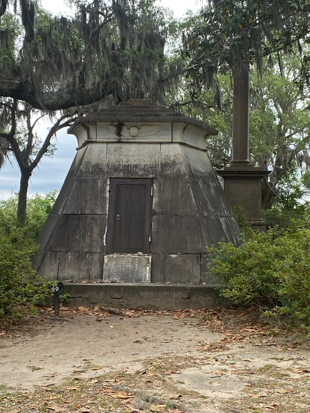 The image shows an aged weathered mausoleum with a pyramid-shaped roof set in a serene environment draped with Spanish moss hanging from surrounding trees