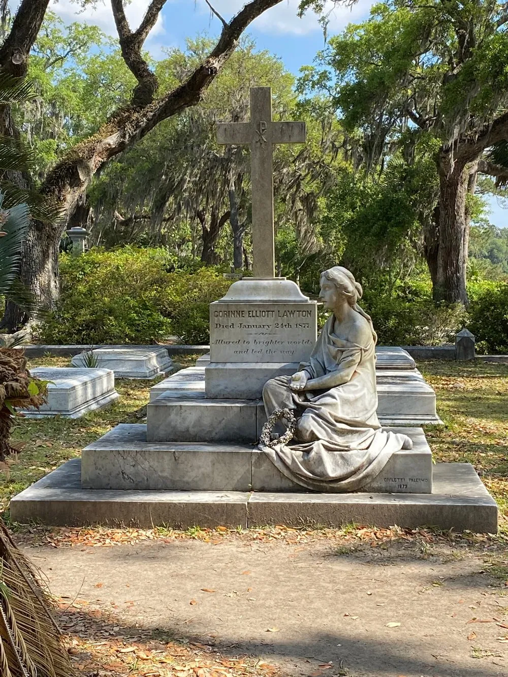 The image shows an elaborate grave with a seated statue of a woman looking contemplatively at a cross surrounded by trees draped with Spanish moss indicative of a serene Southern cemetery