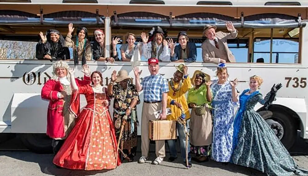 A group of people dressed in various historical and character costumes are smiling and waving in front of a vintage-style trolley bus