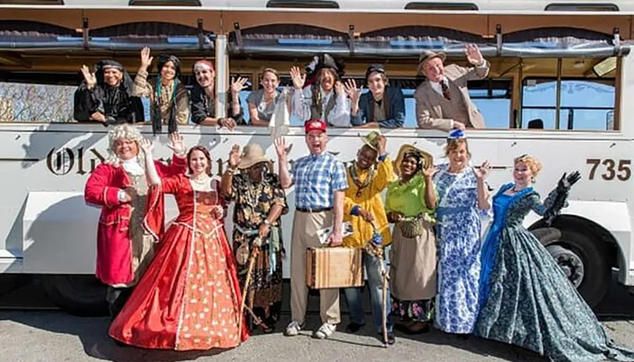 A group of people dressed in various historical and character costumes are smiling and waving in front of a vintage-style trolley bus.