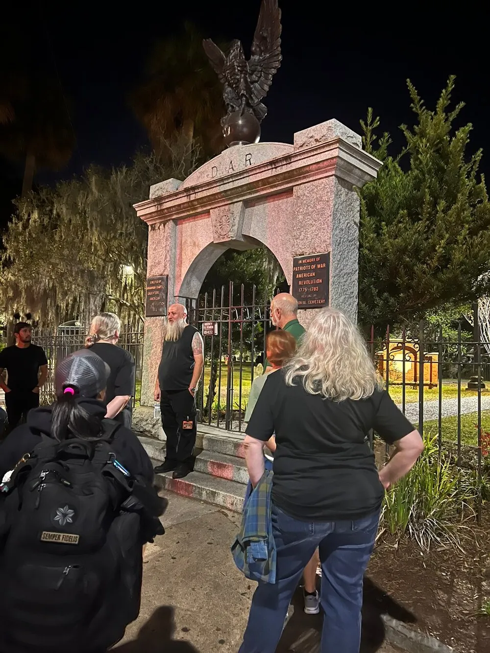 A group of people is gathered at night around a historic monument with the inscription DAR and an eagle statue on top commemorating the patriots of the American Revolutionary War from 1775-1783