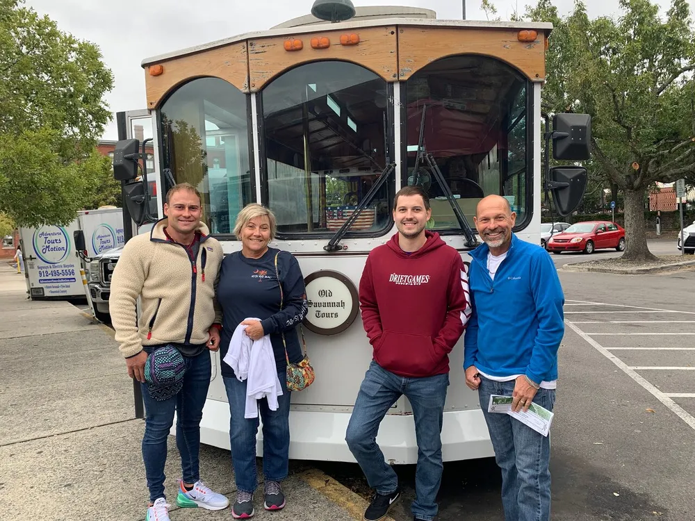 Four people are standing and smiling in front of a white trolley bus perhaps preparing for or returning from a guided tour
