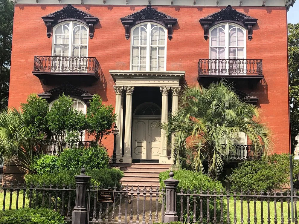 The image shows a well-maintained two-story red brick building with black decorative window accents and balconies surrounded by lush greenery and a wrought-iron fence