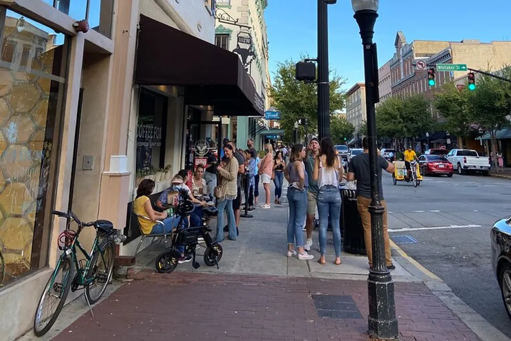 People are gathered on a city sidewalk some seated at an outdoor caf and others standing in a line on a sunny day with cars and buildings in the background