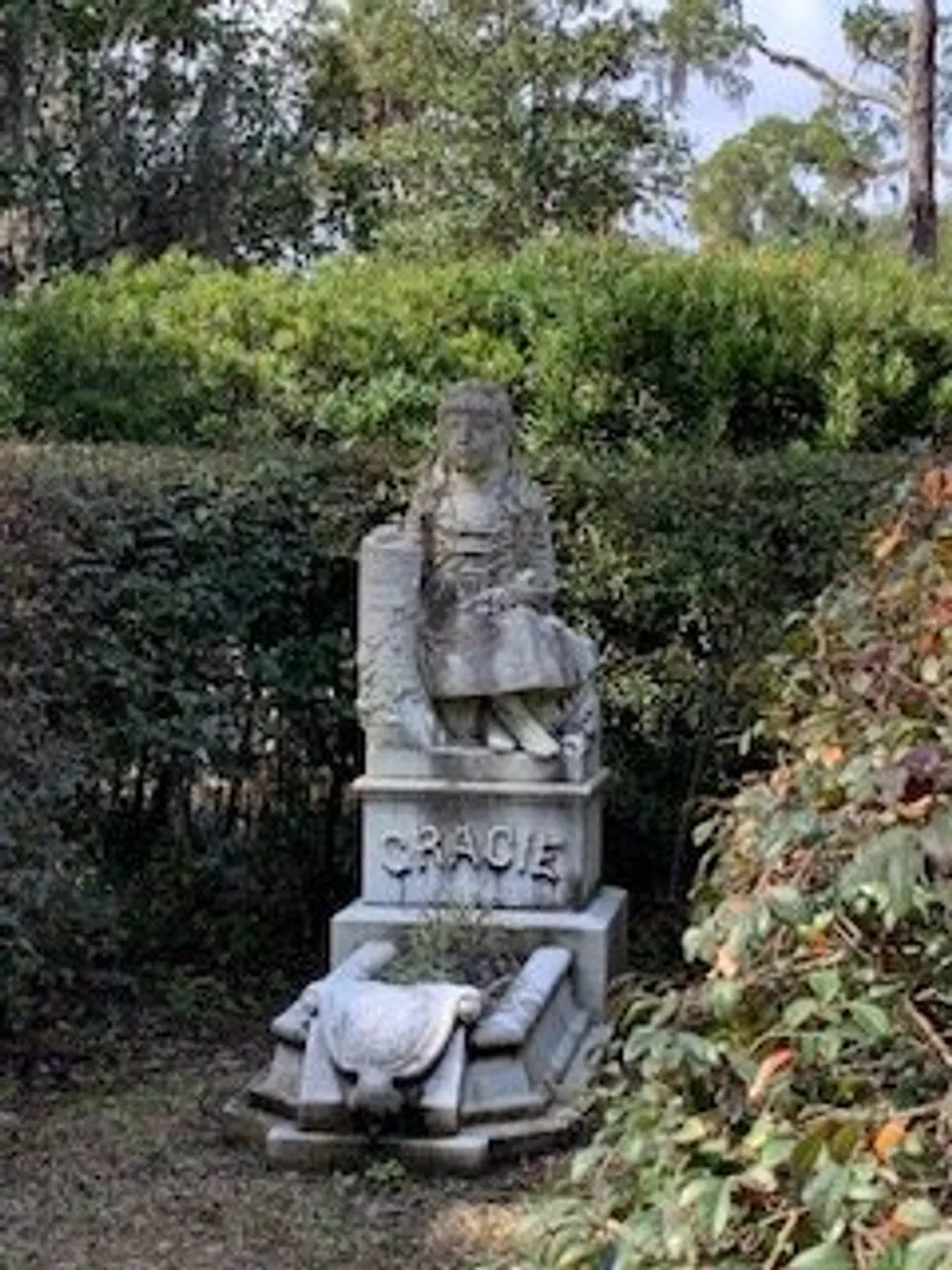 The image shows a weathered stone monument of a small dog sitting atop a pedestal with the name Gracie inscribed on it set within a peaceful green garden environment