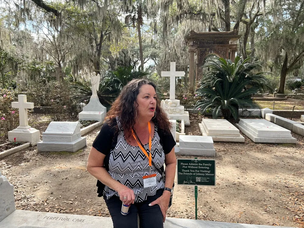 A woman wearing a badge stands in a cemetery adorned with white cross tombstones and Spanish moss-draped trees