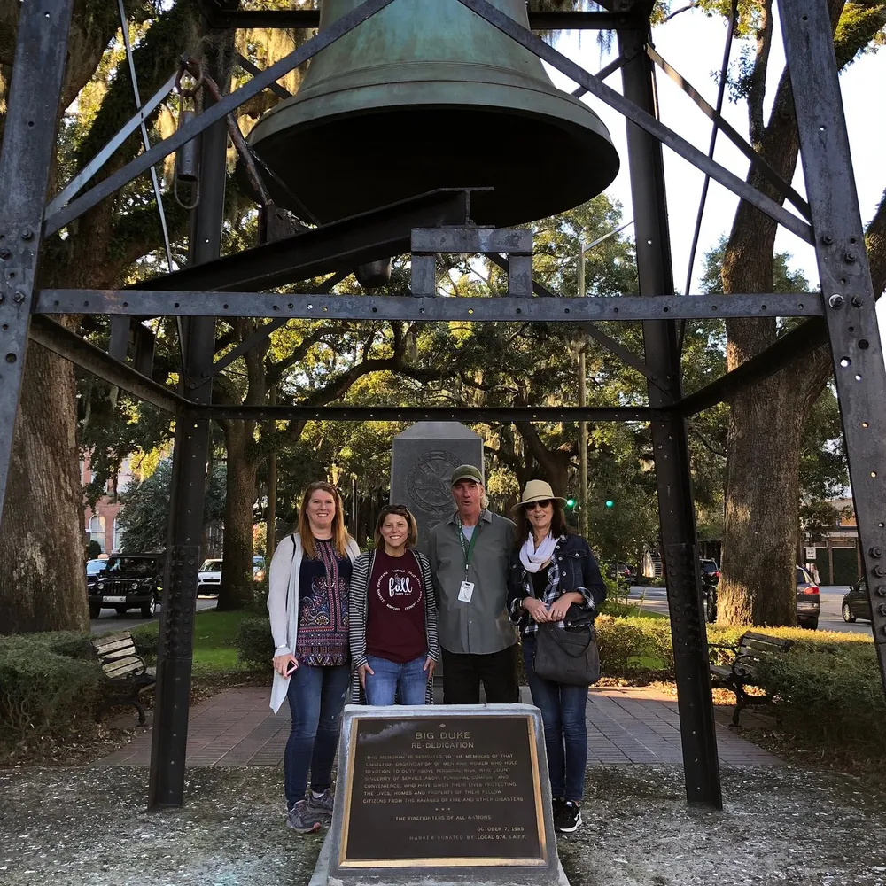 Four individuals are posing for a photo in front of a large historic bell mounted on a metal structure with trees in the background and a plaque titled BIG DUKE at the forefront