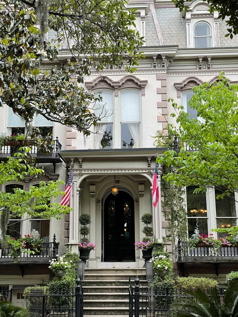 This image shows the facade of an elegant Victorian-style house with an ornate entryway draped with American flags and adorned with flowering planters