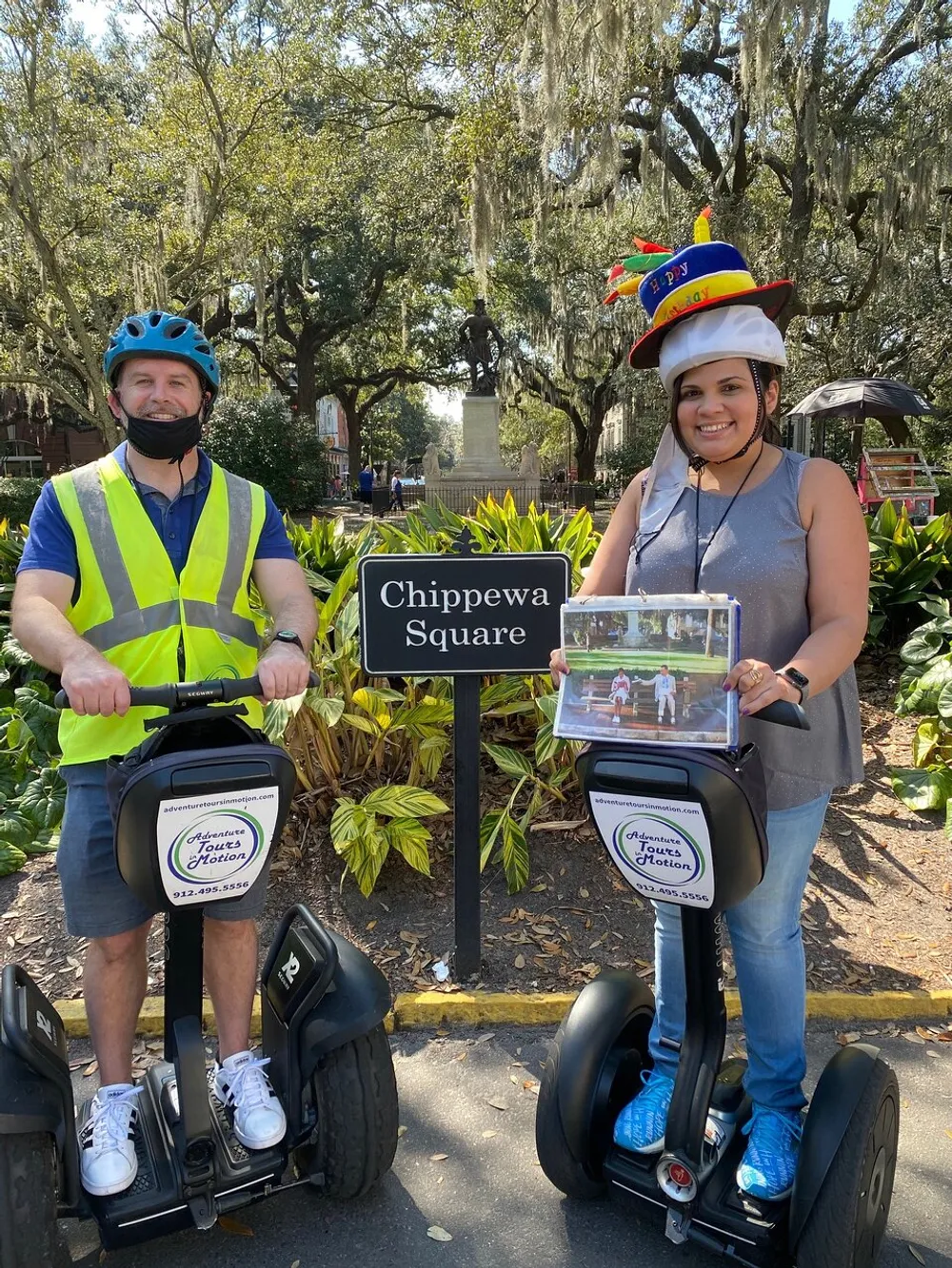 Two individuals are smiling for a photo while standing on Segways at Chippewa Square with the woman wearing a festive hat and holding a sign and the man wearing a safety vest and helmet