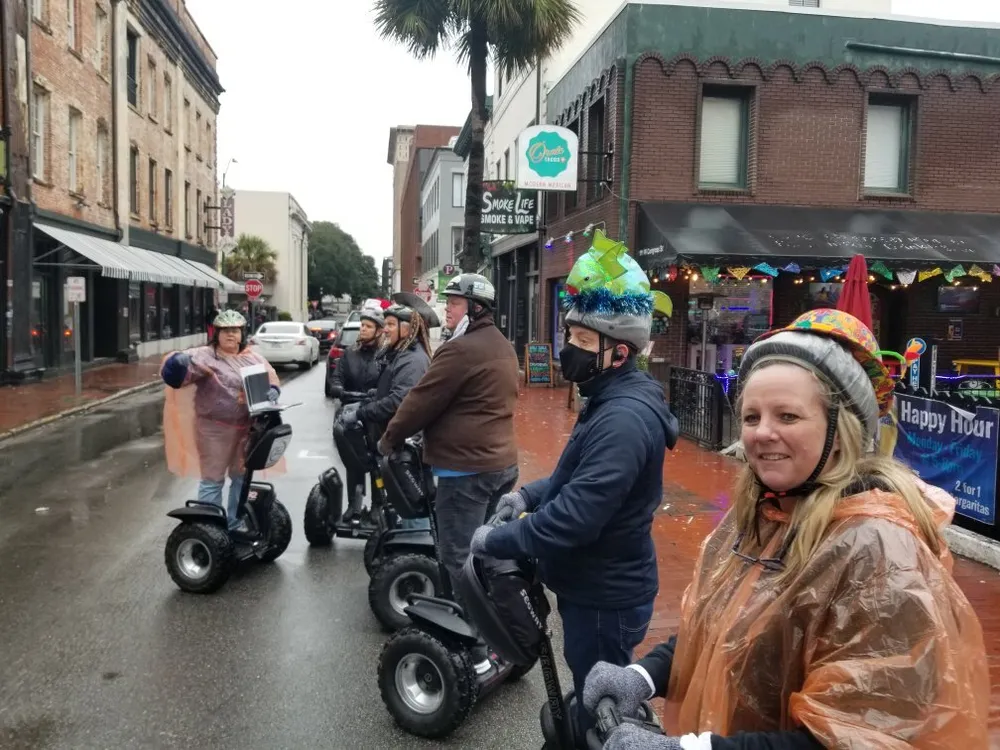 A group of people wearing party hats are riding Segways on a damp city street with one person in the front wearing a poncho and gesturing likely leading a tour