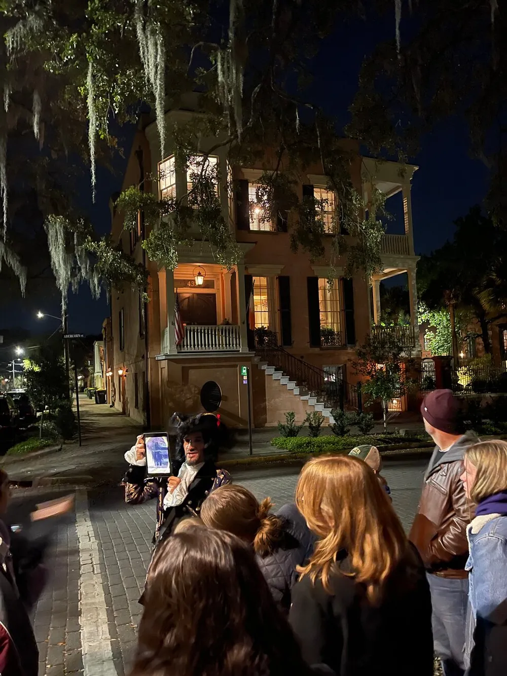 A group of people listen to a costumed guide during a nighttime walking tour in front of an illuminated historic building adorned with Spanish moss