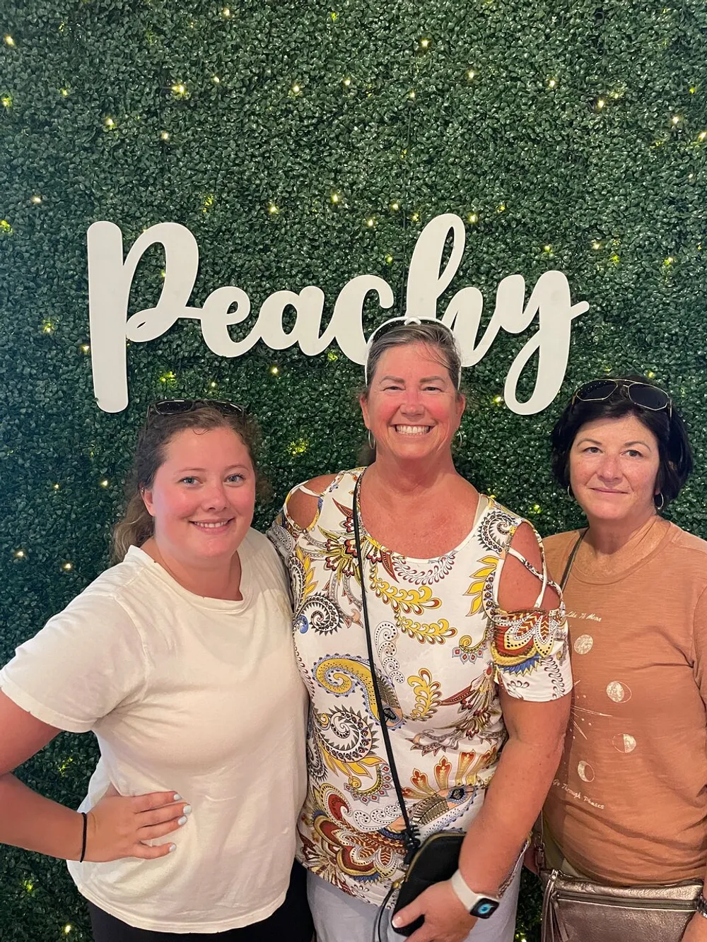Three smiling women are posing together in front of a leafy green wall with the word Peachy written on it
