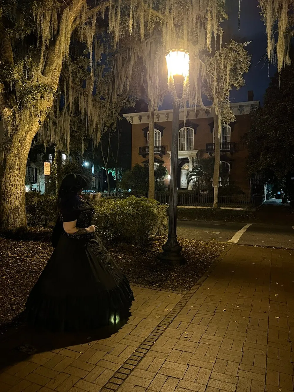 A person in a Victorian-style dress stands under a street lamp at night surrounded by Spanish moss-draped trees
