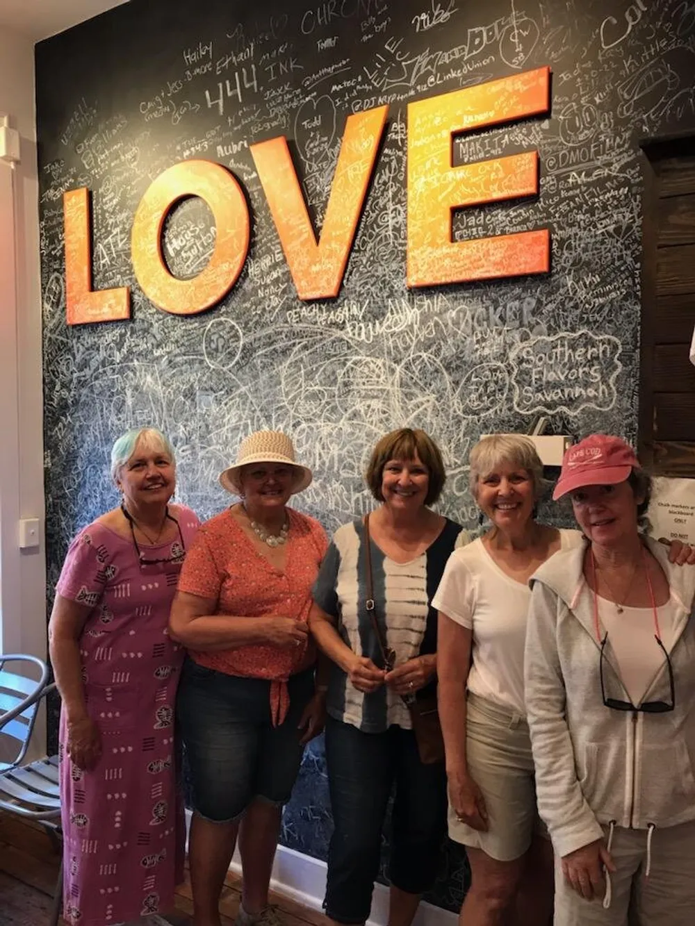 Five smiling women stand in front of a wall with a large illuminated LOVE sign which is covered in handwritten signatures and messages