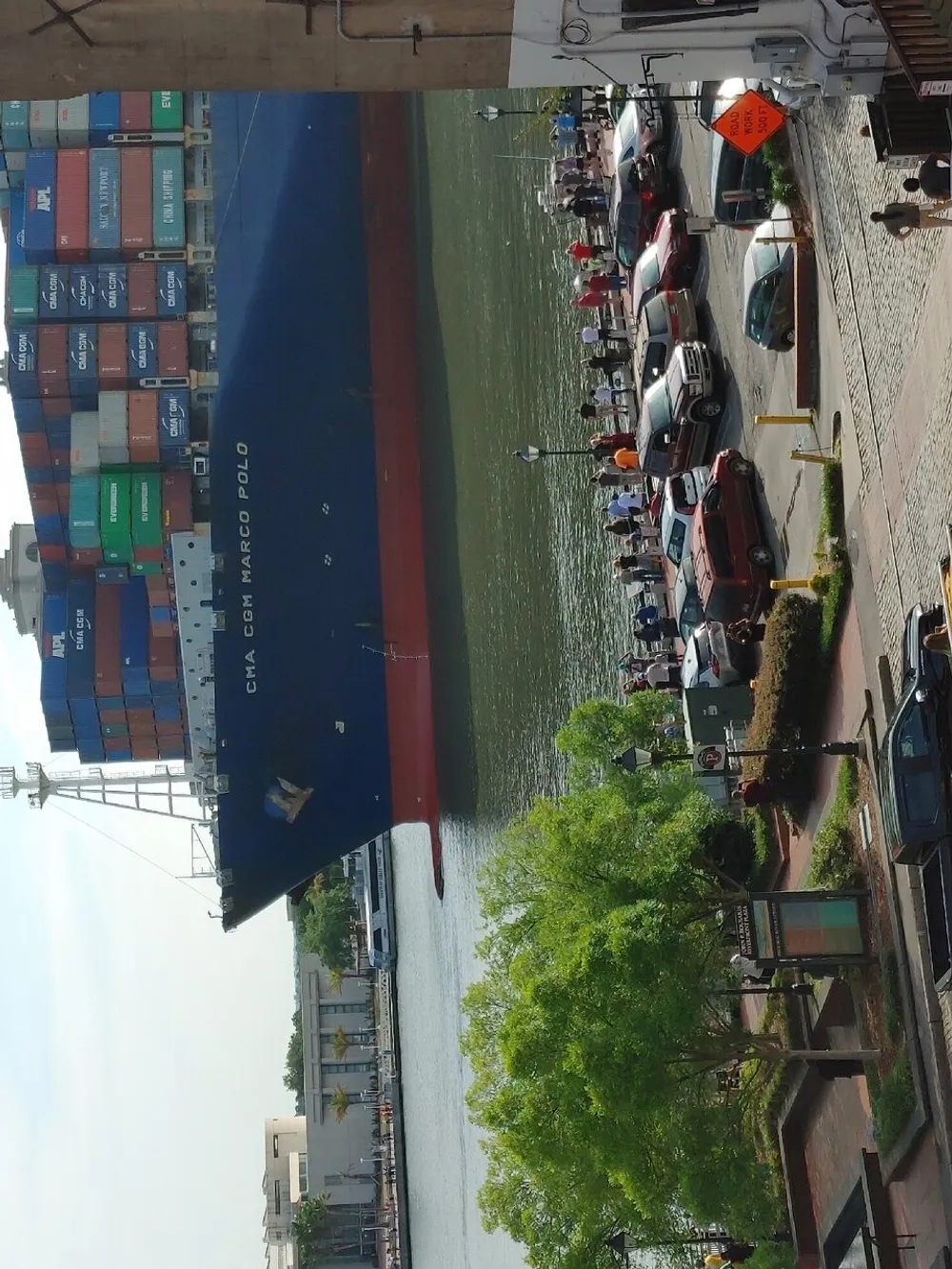 A massive container ship towers over a quayside promenade bustling with people and lined with parked cars