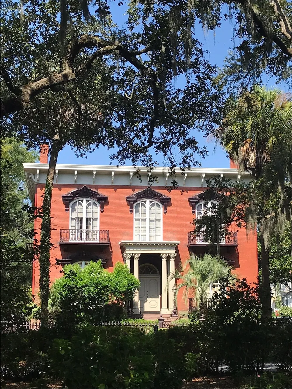 A classic red brick house with white trim balconies and a pillared entrance is nestled amongst lush greenery and trees draped with Spanish moss under a bright blue sky