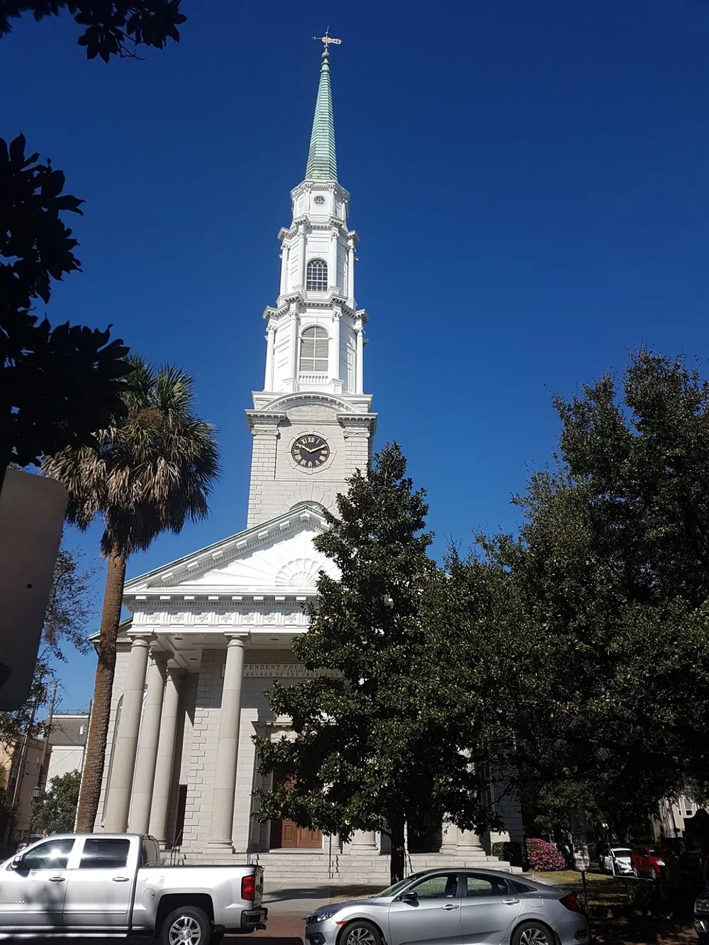 The image shows a tall white church with a steeple and clock under a clear blue sky partially obscured by trees with parked cars along the street in the foreground