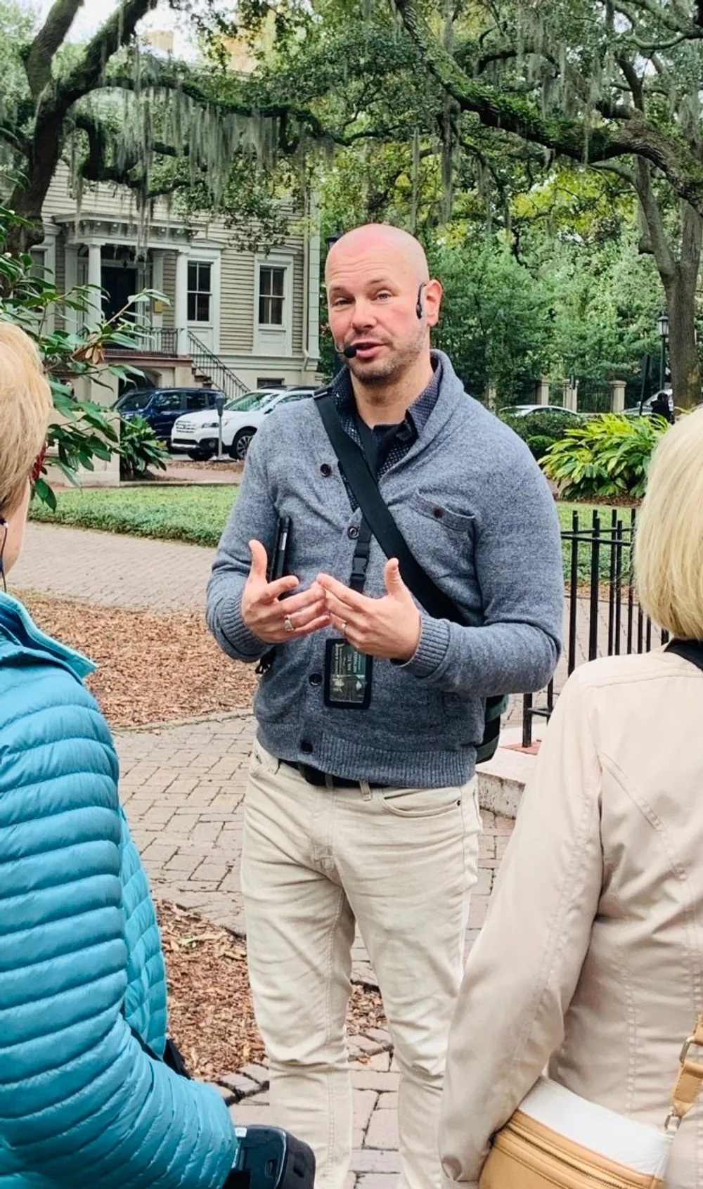 A man with a headset is speaking and gesturing with his hands possibly giving a tour with listeners facing him and a backdrop of trees and historic architecture