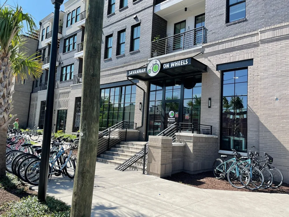 The image shows a sunny day outside a modern brick building that houses a business called Savannah On Wheels with several bicycles parked in racks out front