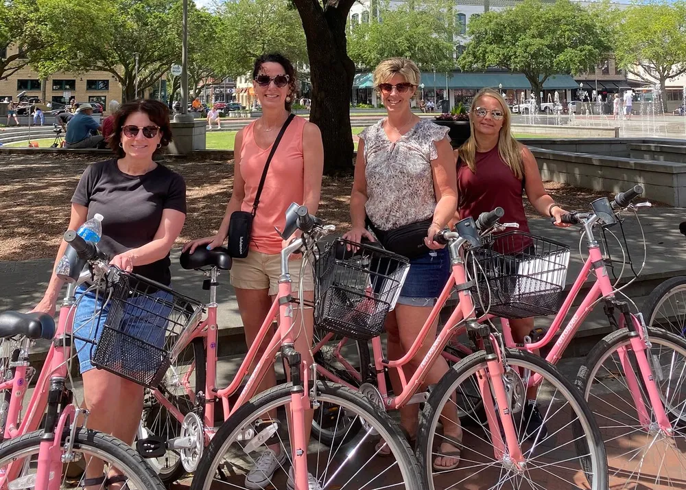 Four women are smiling and standing behind a row of pink bicycles in a sunny outdoor setting