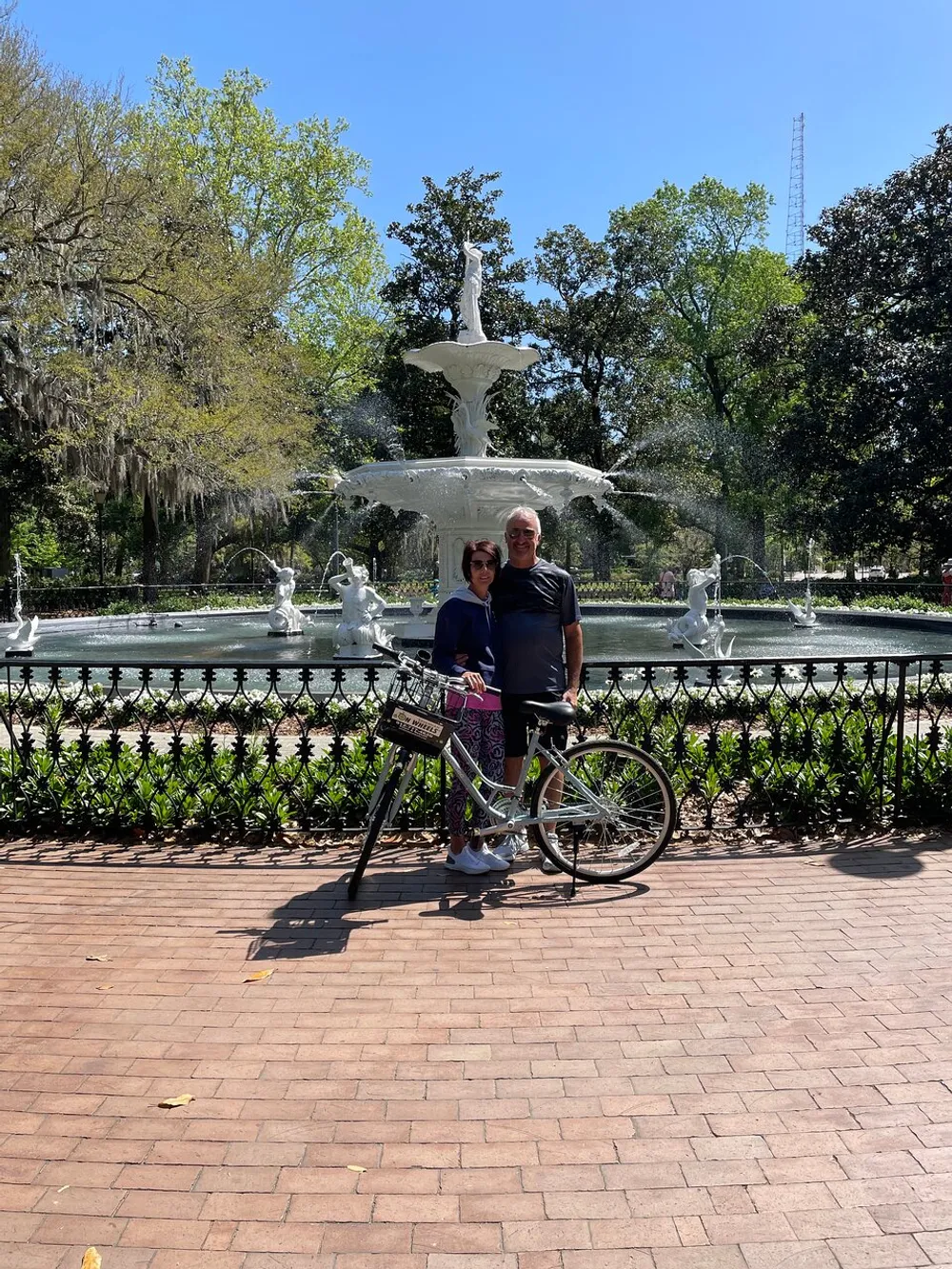Two people are standing in front of a decorative fountain with a bicycle smiling for a photo on a sunny day