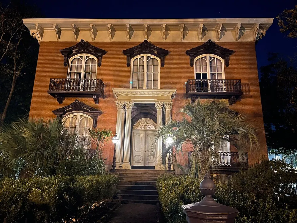 The image shows a stately two-story brick building at night illuminated by warm lights featuring classic architectural details balconies and surrounded by lush greenery
