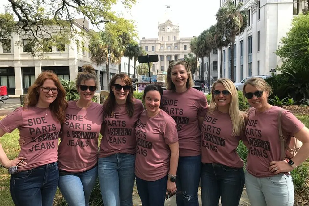 A group of seven smiling women wearing matching pink t-shirts that read SIDE PARTS  SKINNY JEANS pose together outdoors