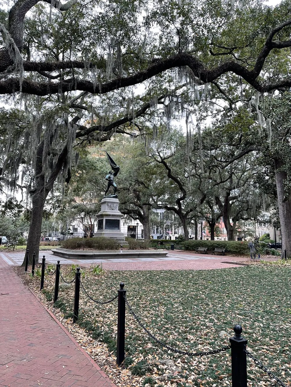 An autumnal park scene featuring a statue atop a pedestal surrounded by leaf-strewn grass live oak trees draped with Spanish moss and a brick pathway