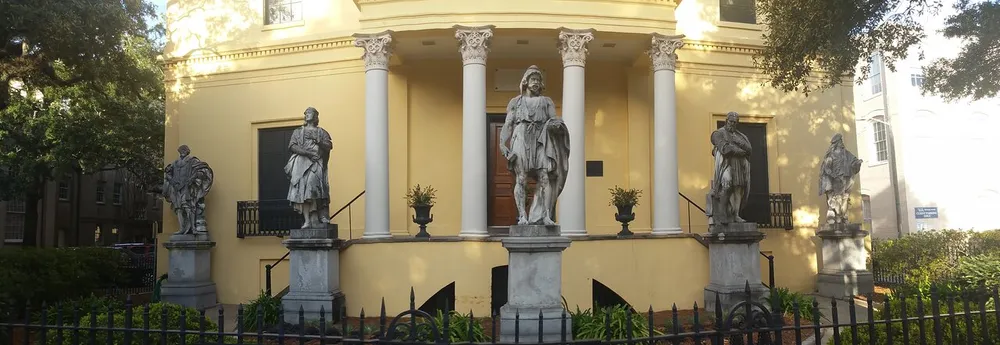 The image shows a yellow classical building adorned with columns and fronted by four statues of historical figures on pedestals
