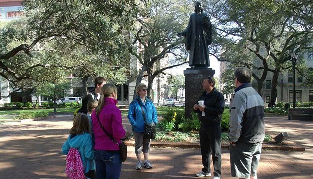 A group of people are gathered around a tour guide next to a statue in a park with lush green trees